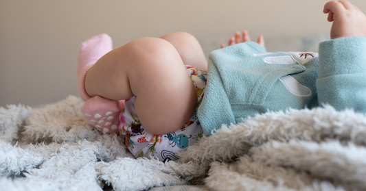 A close-up of a baby lying on a fluffy white blanket, wearing a white cloth diaper with a colorful cartoon print, pink socks, and a light blue fleece top.